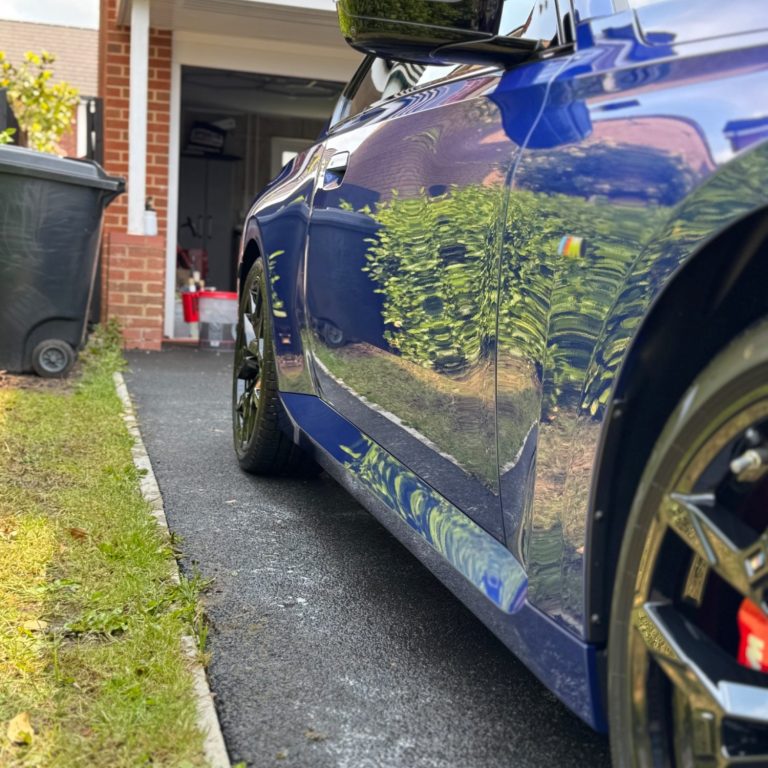 Close-up of a blue car parked on a driveway with a green tree reflection on the side.