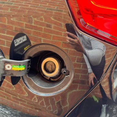 Fuel cap on a car with a reflection of a person and brick wall in the background.